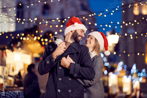 Christmas spouses hugging on the street on New Year. A young happy couple in love with Santa hats on their heads is standing on the street, hugging and looking at each other on New Year's eve.