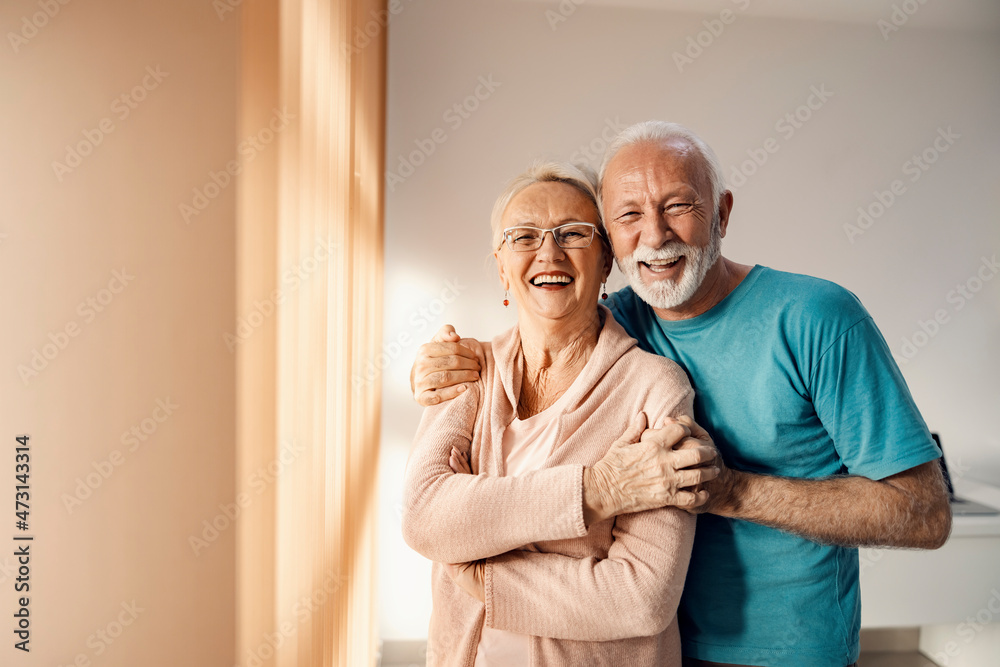Senior couple hugging in a nursing home. A happy senior couple standing next to a window in a nursing home, hugging and smiling. They have all care they need. - obrazy, fototapety, plakaty 