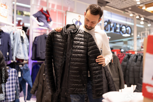 Adult handsome brunet unshaven man chooses warm black jacket in a clothing store in a shopping mall. Outerwear and shopping