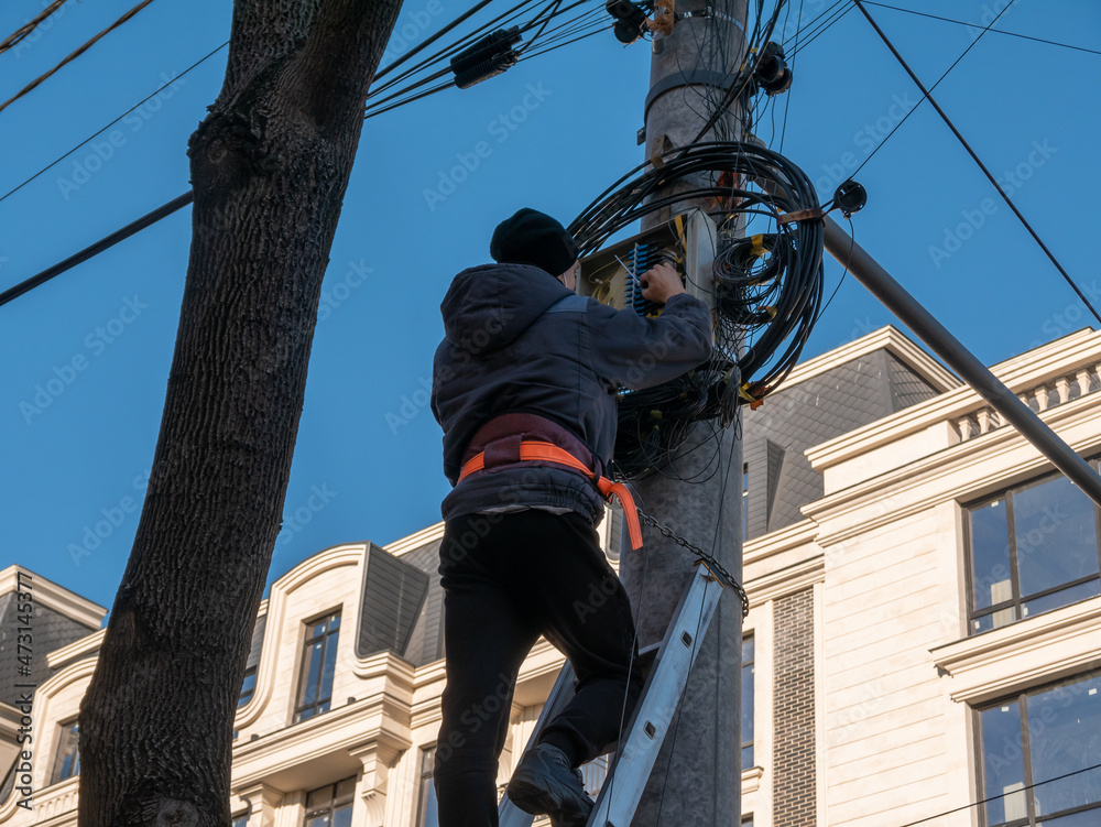An electrician on a ladder installs a junction box for the Internet on a pole.