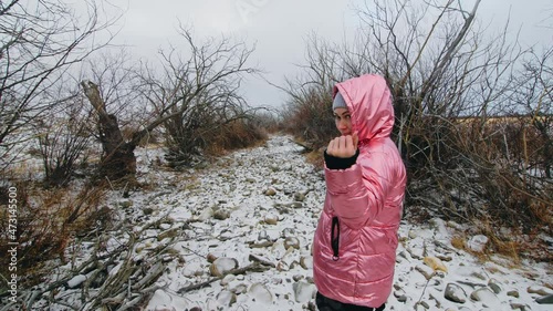 Young woman in a winter pink down jacket with a hood walks in the forest in winter. Dry mouth of the river, dark magical view. photo