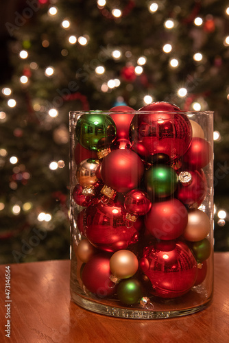Holiday table center piece of a glass jar filled with Christmas ornaments and a lit tree in the background.