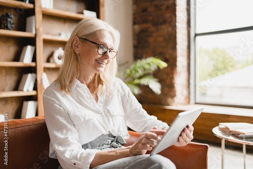 Caucasian middle-aged mature businesswoman teacher tutor freelancer noting new ideas, business plans in digital tablet, working remotely in office, searching data in social media online
