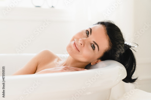 Naked caucasian mature middle-aged woman relaxing while taking shower bath in hot warm water at home looking at camera