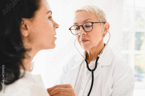 Serious caucasian female family doctor general practitioner listening in stethoscope female mature patient s chest  heart beat at hospital appointment