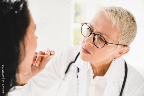 Caucasian mature senior female family doctor general practitioner checking examining attentively female patient`s throat with medicine stick. Angina flu prevention photo