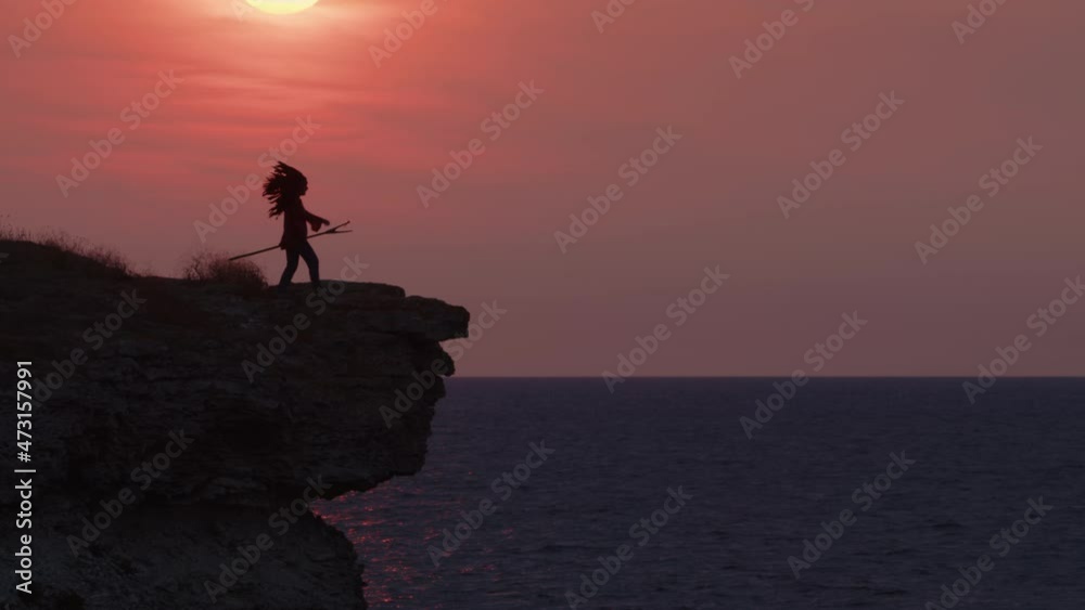 Silhouette of Native American in headdress with feathers approaches edge of cliff of sea coast and looks into distance against background of red sunset sky. Silhouette of Native American in headdress with feathers approaches edge of cliff of sea coast and looks into distance against background of red sunset sky.