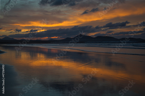Sunset at Famara Beach. Mountains in the background and orange clouds reflecting in the wet sand. Lanzarote, Spain.