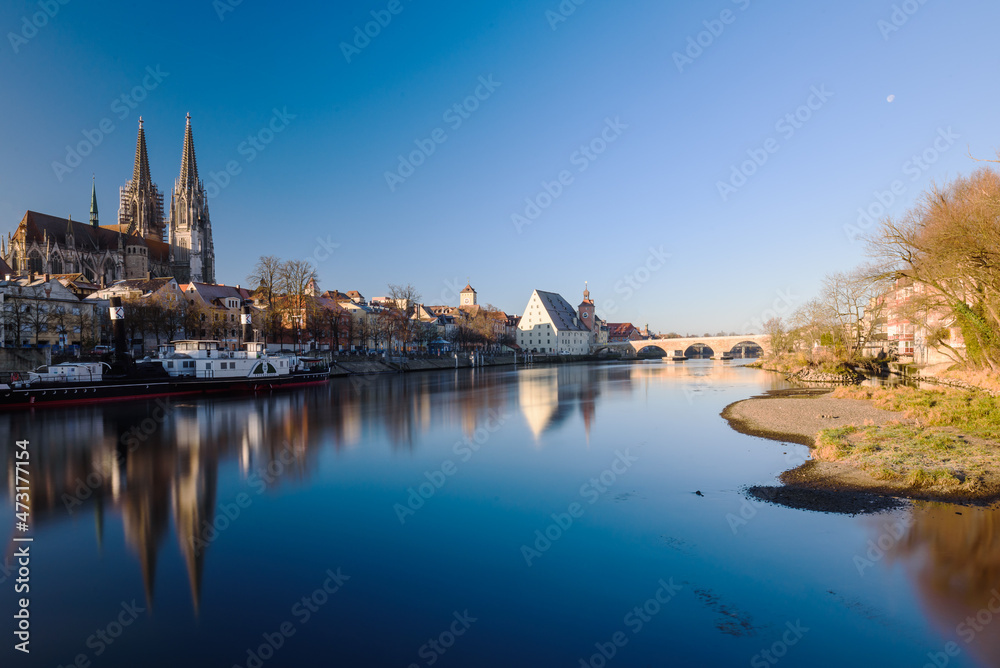 Donauufer in Regensburg mit Dom, Spiegelungen und steinerner Brücke bei blauem Himmel,  im Abendlicht im Winter