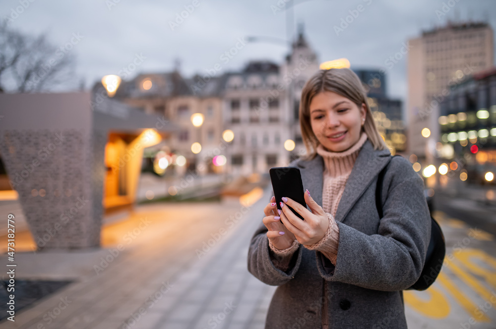 Beautiful charming girl in coat standing in street and looking at telephone.Nice winter day.