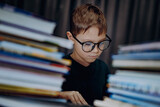 cute caucasian boy wearing glasses covering head with book. Cheerful boy peeking from behind piles of books
