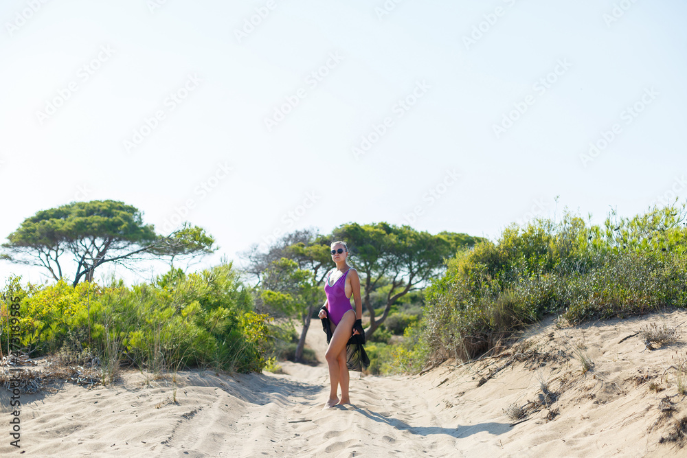 Naklejka premium A slender tanned young woman in a swimsuit and a pareo, standing on the golden sand in the desert. Model woman resting and sunbathing on vacation. Travel concept, swimwear