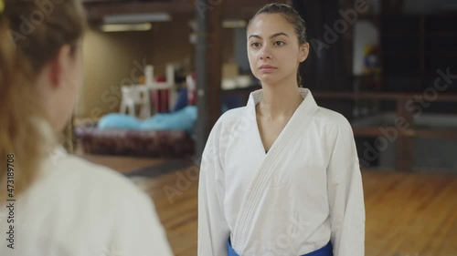 Medium shot of female fighters bowing before karate training. Front view of confident Caucasian girl in kimono standing in gym, showing respect for opponent by bowing. Greeting, martial arts concept