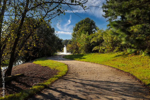 a shot of a long smooth winding footpath near a like with a water fountain surrounded by lush green and autumn colored trees and plants with blue sky and clouds at Garrard Landing Park in Alpharetta photo