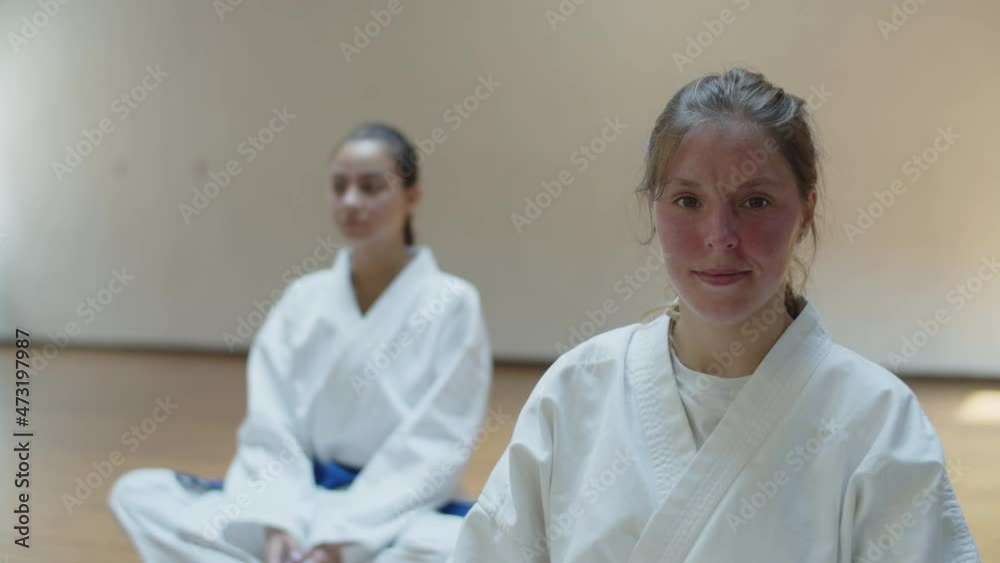 Front view of cheerful martial artsist sitting on floor in gym. Medium shot of beautiful Caucasian girl wearing white kimono, looking at camera while relaxing after training. Sport, emotion concept Front view of cheerful martial artsist sitting on floor in gym. Medium shot of beautiful Caucasian girl wearing white kimono, looking at camera while relaxing after training. Sport, emotion concept