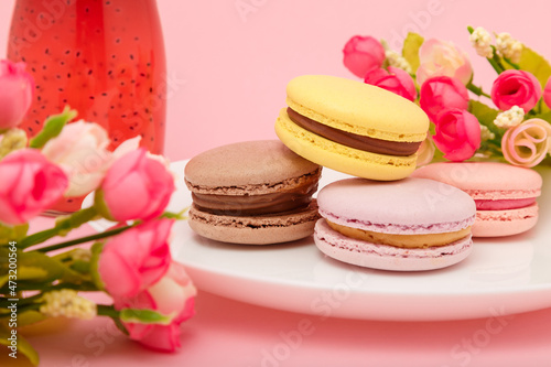 Plate of macaroons on pink background with flowers. Drink in bottle. Sweet pastry, baked products, sweets, dessert. Unhealthy diet.