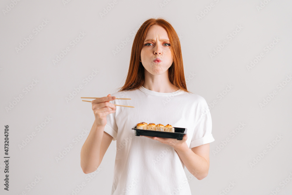 Studio portrait of hungry young woman eating delicious sushi rolls with chopsticks looking at camera standing on white isolated background. Pretty Caucasian redhead female chewing Asian food. Studio portrait of hungry young woman eating delicious sushi rolls with chopsticks looking at camera standing on white isolated background. Pretty Caucasian redhead female chewing Asian food.