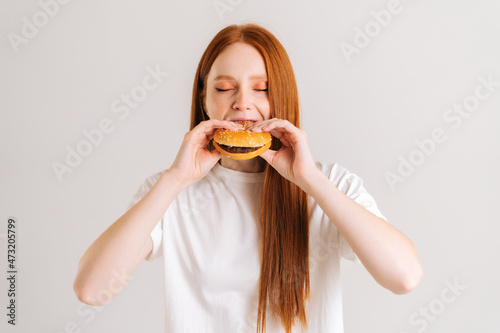 Studio shot of attractive young woman with closed eyes enjoying bite of appetizing delicious hamburger on white isolated background. Closeup front view of female eating tasty burger .