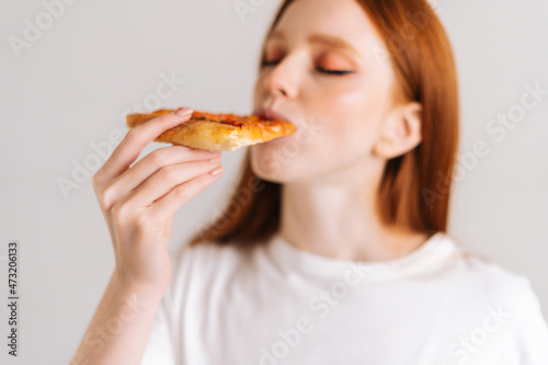 Close-up face of happy attractive young woman with closed eyes appetite eating delicious pizza standing on white isolated background. Pretty redhead female eating tasty meal  selective focus.