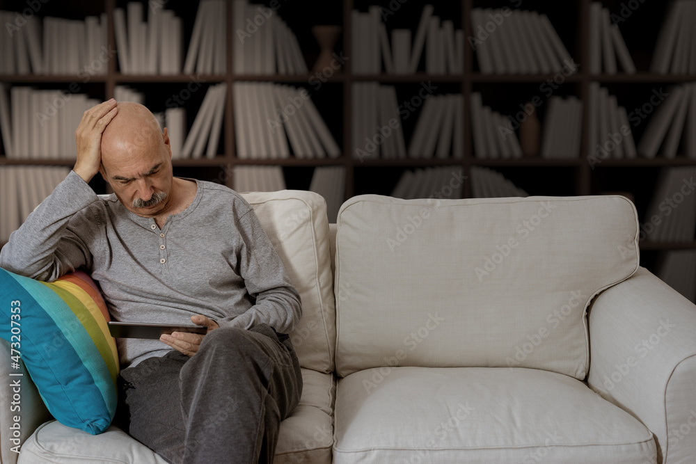 Obraz premium Senior Male Reading in a Digital Tablet Sitting at the Sofa at the House Living Room