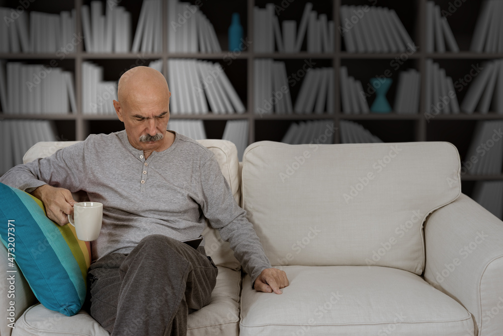 Senior Male Reading in a Digital Tablet Sitting at the Sofa at the House Living Room Senior Male Reading in a Digital Tablet Sitting at the Sofa at the House Living Room