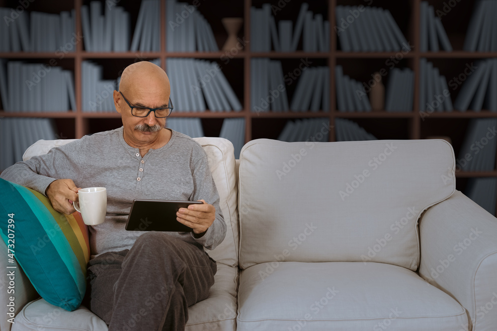 Senior Male Reading in a Digital Tablet Sitting at the Sofa at the House Living Room Senior Male Reading in a Digital Tablet Sitting at the Sofa at the House Living Room
