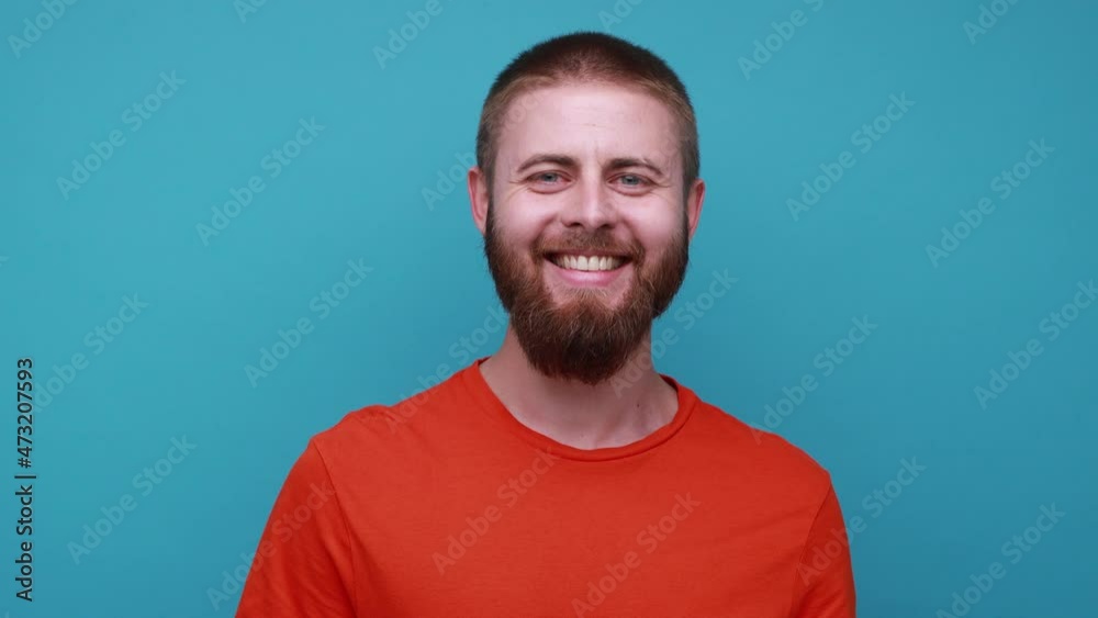 Happy satisfied delighted bearded man standing looking at camera with optimistic expression, being in good mood, wearing orange T-shirt. Indoor studio shot isolated on blue background. Happy satisfied delighted bearded man standing looking at camera with optimistic expression, being in good mood, wearing orange T-shirt. Indoor studio shot isolated on blue background.