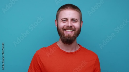 Happy satisfied delighted bearded man standing looking at camera with optimistic expression, being in good mood, wearing orange T-shirt. Indoor studio shot isolated on blue background.