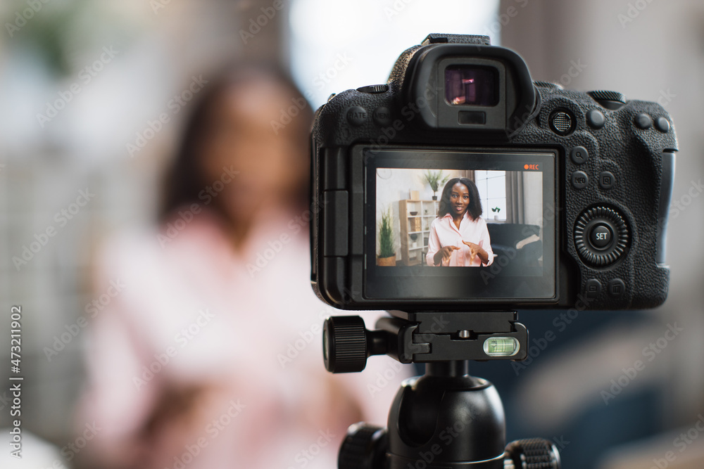 Close up of digital camera with positive african american woman on screen. Blur background of female influencer gesturing and talking while sitting at desk. Blogging concept.