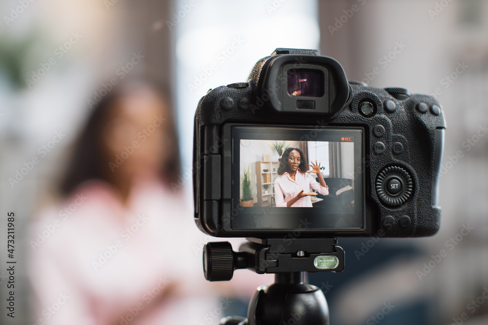 Blur background of african american woman in eyeglasses and pink shirt recording new video blog for her social networks. Focus on screen of digital camera.