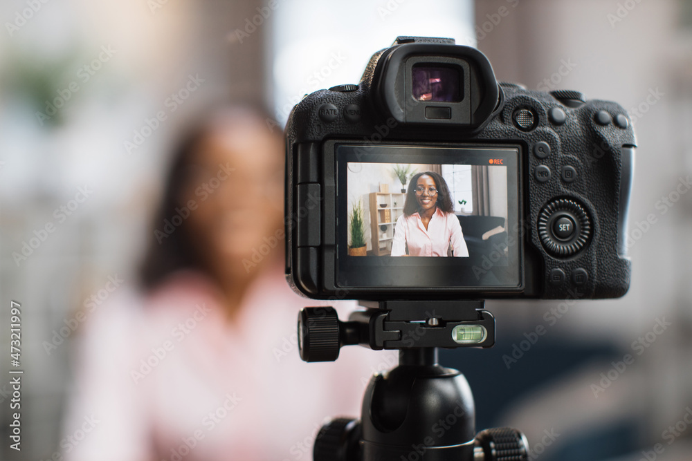 Smiling african woman talking and gesturing in front of modern video camera while sitting on blur background. Live streaming concept.