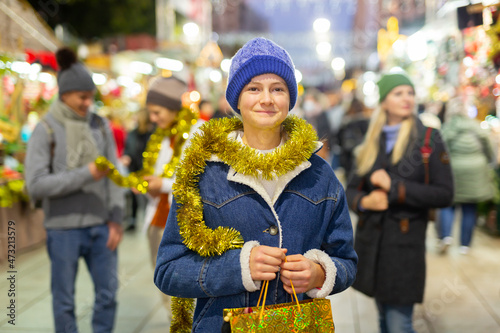 Positive teenager girl standing at christmas fair and holding bag with gift.