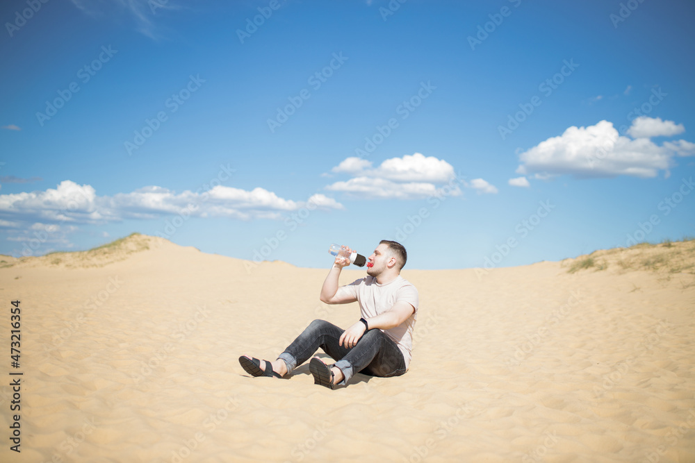 Young tired man drinking water while sitting on the top of sand dune in desert, sand is al around and some greens. Hot summer weather, concept of travelling around planet. Great Europen desert Young tired man drinking water while sitting on the top of sand dune in desert, sand is al around and some greens. Hot summer weather, concept of travelling around planet. Great Europen desert