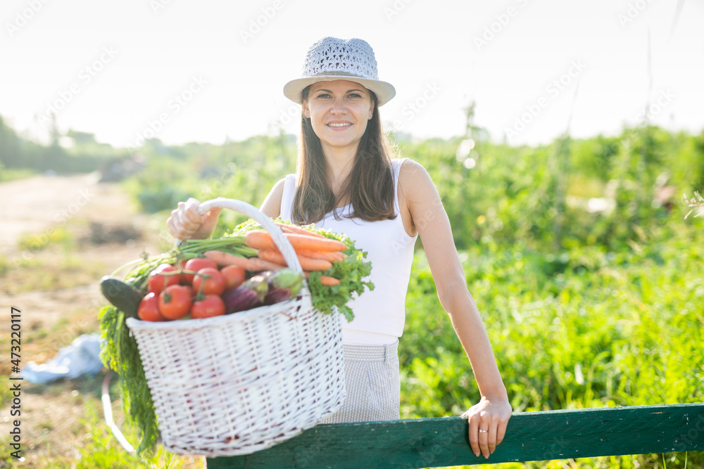 Fototapeta premium Happy smiling woman with basket of ripe vegetables in the summer garden
