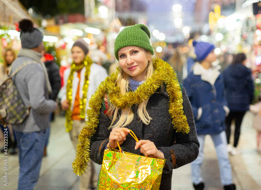 Positive woman standing at christmas fair and holding bag with gift.