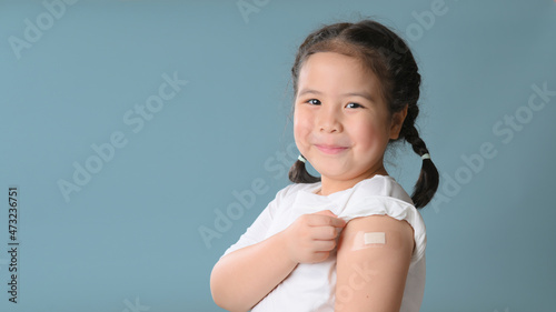 Coronavirus Vaccination Advertisement. Happy Vaccinated Little asian girl Showing Arm With Plaster Bandage After Covid-19 Vaccine Injection Posing Over Blue Background, Smiling To Camera. Blank Space