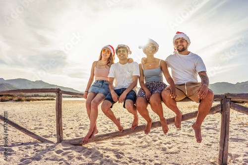 Suggestive photography with backlight sun effect of four young happy people enjoying outdoor life in exotic vacation resort sitting on wooden fence on a white sand beach at sunset