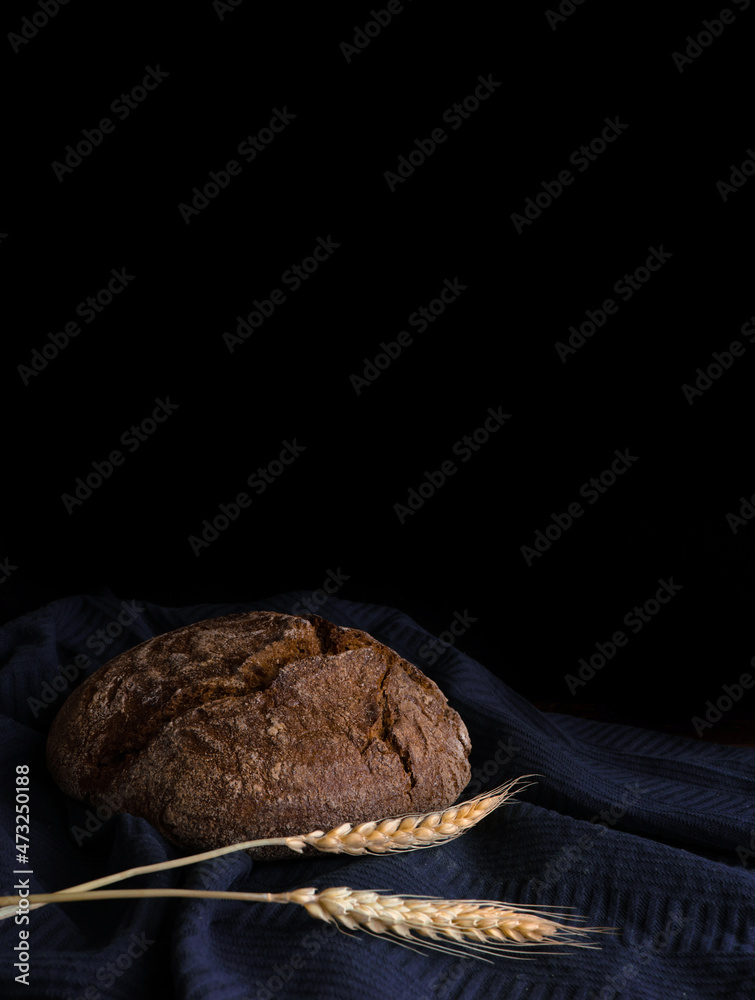 Round loaf of rye bread with a spike of wheat on a blue towel in a dark key on a black background, vertical photo, free space for text Round loaf of rye bread with a spike of wheat on a blue towel in a dark key on a black background, vertical photo, free space for text