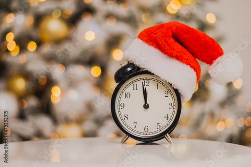 Obraz na plátne Alarm clock in a santa hat against the background of a christmas tree