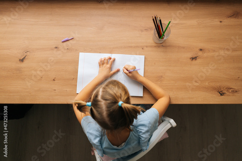High-angle view of unrecognizable primary little child girl doing homework sitting at home table by window. Smart preschool kid studying alone drawing with pen in bedroom. Homeschooling concept.