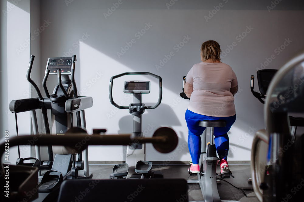 Rear view of mid adult overweight woman exercising on stationary bike indoors in gym