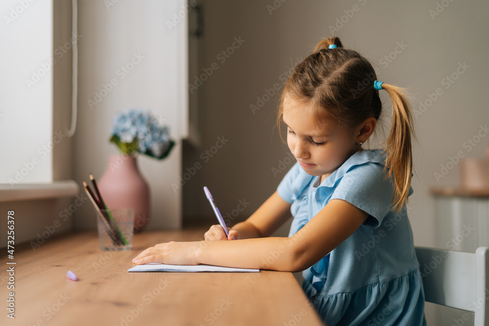 Side view of focused primary little child girl learning writing doing homework sitting at home table by window. Portrait of smart preschool kid studying alone making note with pen at bedroom. Side view of focused primary little child girl learning writing doing homework sitting at home table by window. Portrait of smart preschool kid studying alone making note with pen at bedroom.