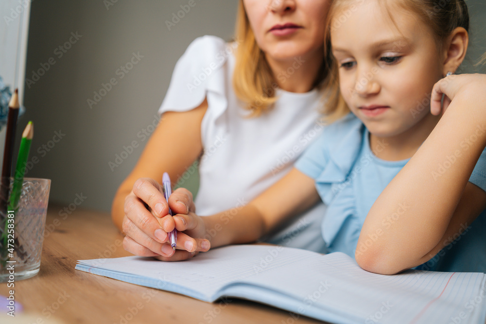 Close-up cropped shot of primary daughter doing homework with young mother sitting at home table by window. Pretty female tutor teaching elementary girl helping with lesson, selective focus. Close-up cropped shot of primary daughter doing homework with young mother sitting at home table by window. Pretty female tutor teaching elementary girl helping with lesson, selective focus.