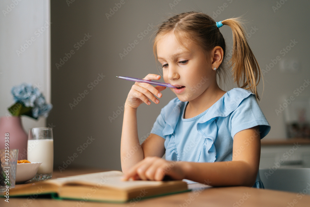Close-up of thinking pretty elementary child school girl doing homework and holding pen against mouth sitting at home table. Pensive preschool kid reading paper book and writing in copybook. Close-up of thinking pretty elementary child school girl doing homework and holding pen against mouth sitting at home table. Pensive preschool kid reading paper book and writing in copybook.