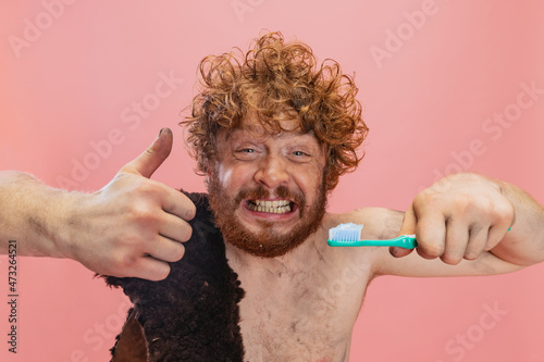 Cropped portrait of funny man in character of neanderthal brushing teeth with toothpaste isolated over pink background. Showing like gesture photo