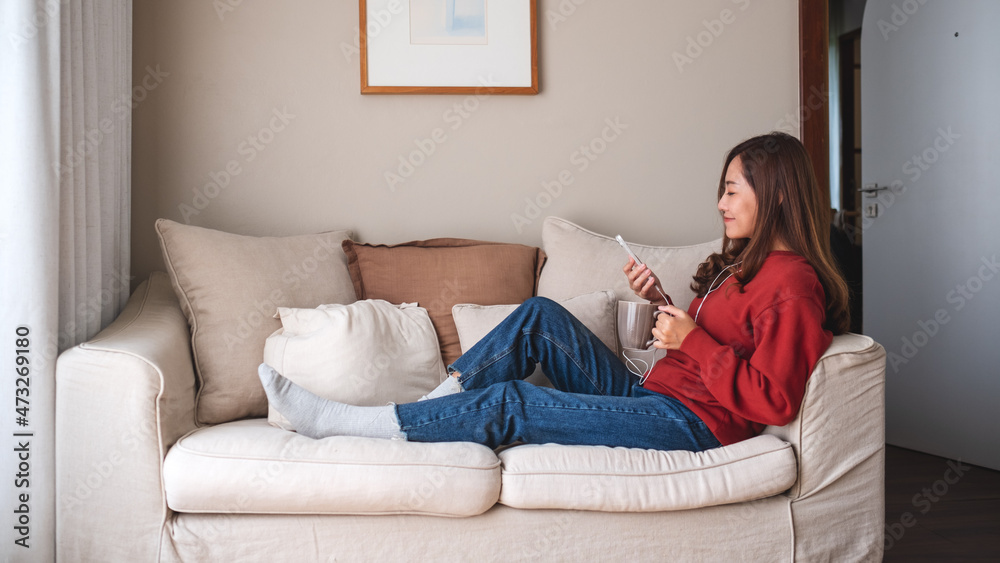 Portrait image of a beautiful young asian woman using mobile phone and earphones to listening to music or online chatting on sofa at home Portrait image of a beautiful young asian woman using mobile phone and earphones to listening to music or online chatting on sofa at home