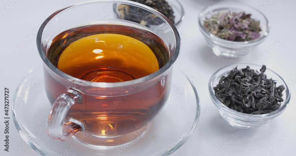 Drop slowly falls into a glass cup of tea. Around there are small bowls with different types of leaf tea. White background. Drop slowly falls into a glass cup of tea. Around there are small bowls with different types of leaf tea. White background.