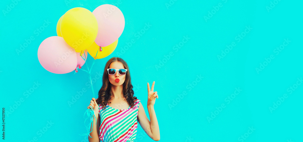 Portrait of beautiful young woman with bunch of balloons blowing her lips sends air kiss wearing a colorful dress on blue background Portrait of beautiful young woman with bunch of balloons blowing her lips sends air kiss wearing a colorful dress on blue background