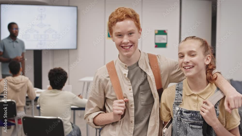 Medium of cheerful Caucasian girl and red-haired boy wearing backpacks, standing in classroom during lesson, talking, smiling and looking on camera Medium of cheerful Caucasian girl and red-haired boy wearing backpacks, standing in classroom during lesson, talking, smiling and looking on camera