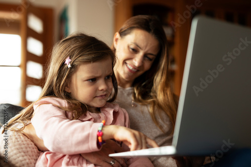 Cheerful little girl using laptop. Beautiful girl watching cartoon with mom. © JustLife